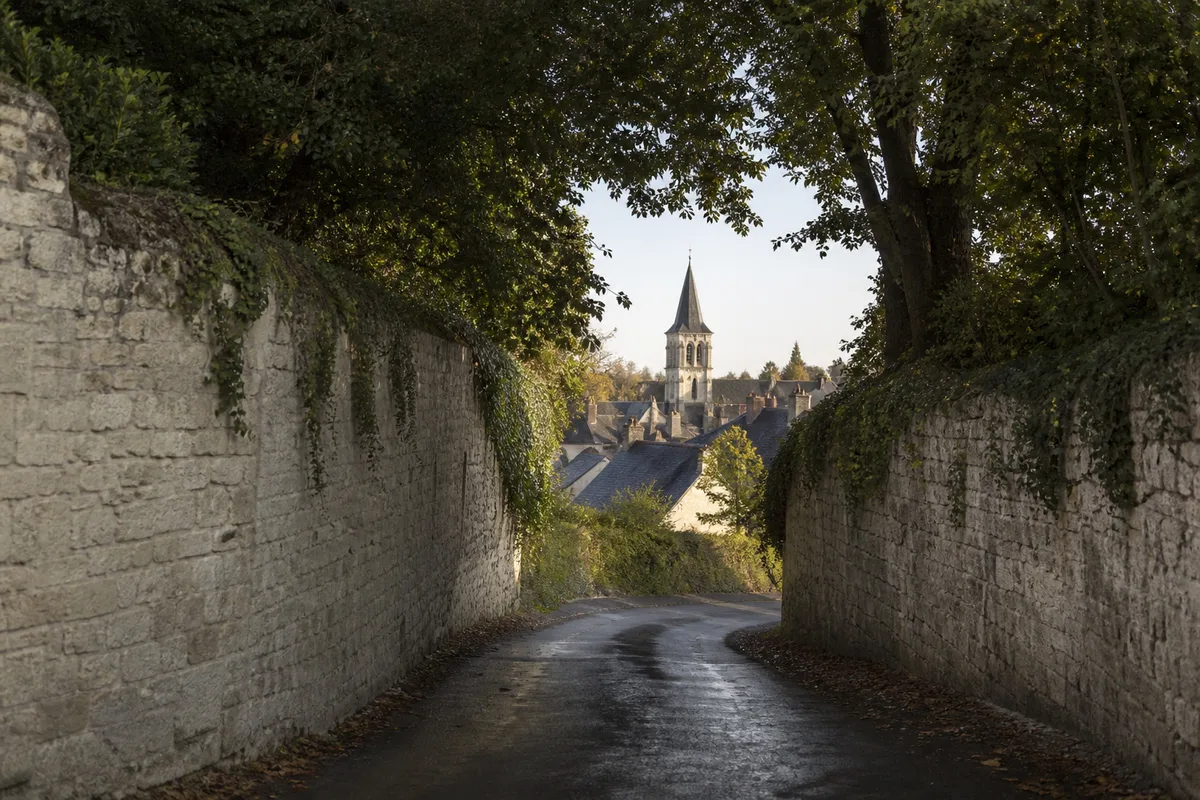 Route départementale étroite du Val de Loire bordée de murs de tuffeau clair, virage ombragé et clocher d’un bourg en arrière-plan sous une lumière matinale